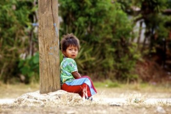 A lone Mayan boy sitting under a basketball hoop on the edge of a village’s playground.